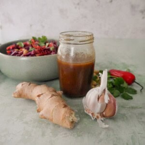 A jar of soy & ginger dressing with fresh root ginger and a bulb of garlic set alongside, and a bowl of peanut and ginger slaw set alongside.
