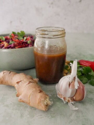 A jar of soy & ginger dressing with fresh root ginger and a bulb of garlic set alongside, and a bowl of peanut and ginger slaw set alongside.