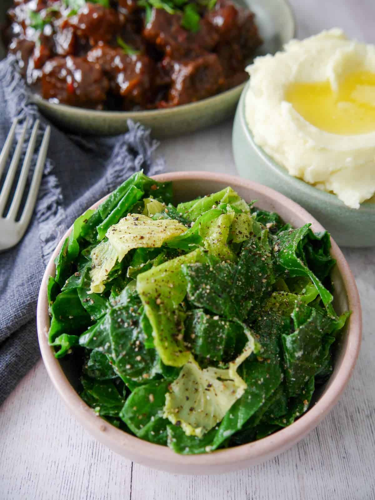 A bowl of buttered spring greens with a bowl paprika beef and a bowl of creamy mashed potato set alongside.