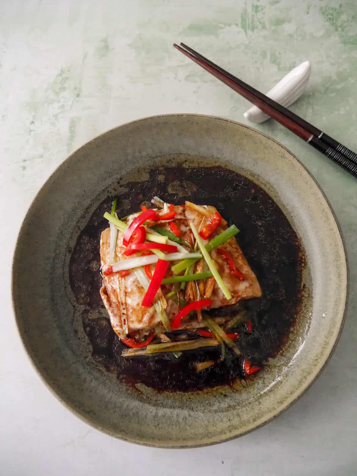 A bowl of Chinese steamed fish fillet with sliced root ginger, spring onion and red chilli, with a hot soy and sesame sauce poured over.
