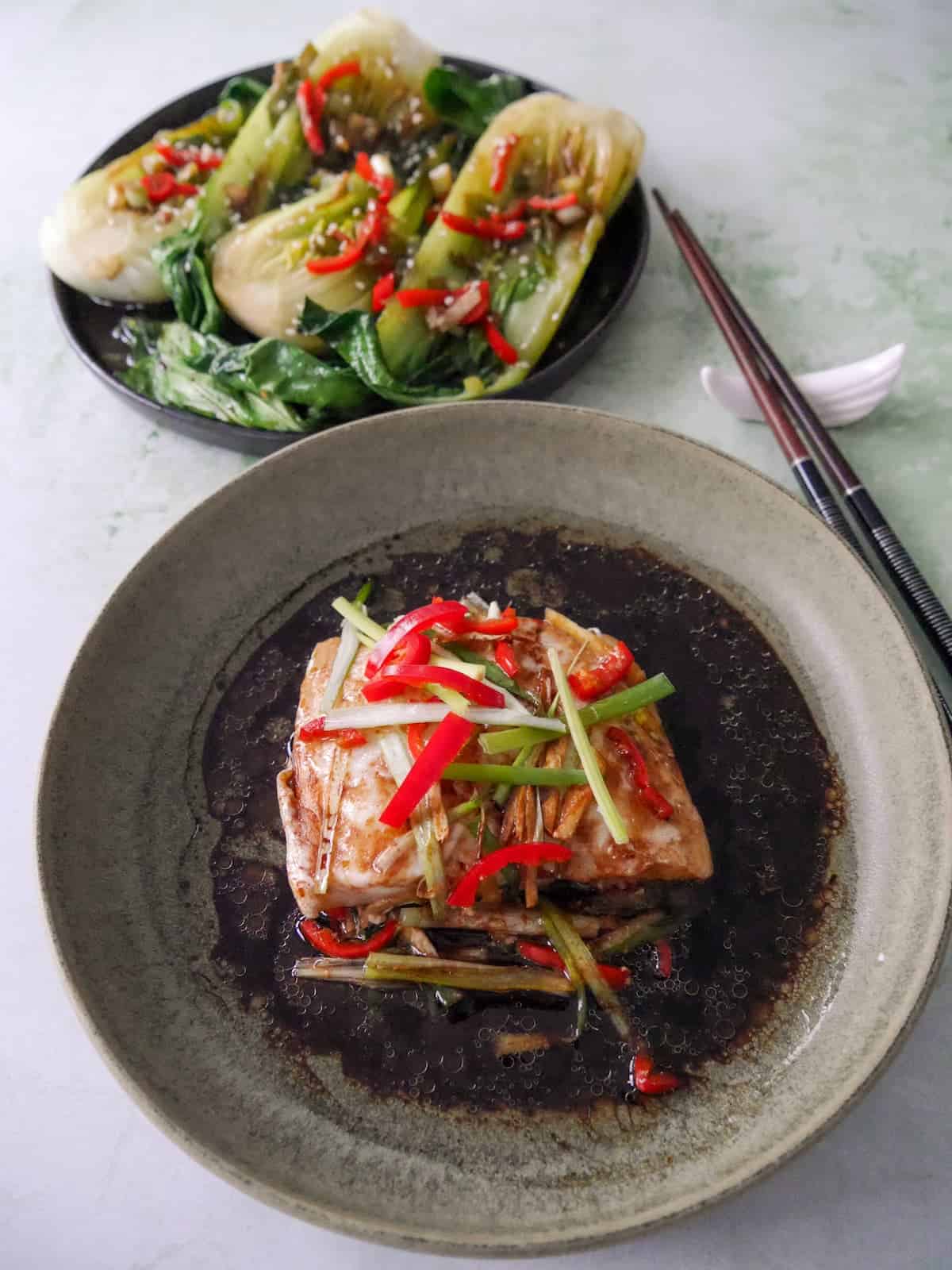 A bowl of Chinese steamed fish fillet with sliced root ginger, spring onion and red chilli, with a hot soy and sesame sauce poured over, and a plate of stir fry pak choi set alongside.