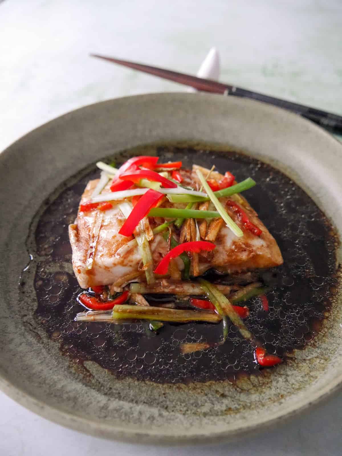 A bowl of Chinese steamed fish fillet with sliced root ginger, spring onion and red chilli, with a hot soy and sesame sauce poured over.