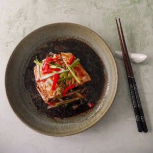 A bowl of Chinese steamed fish fillet with sliced root ginger, spring onion and red chilli, with a hot soy and sesame sauce poured over.