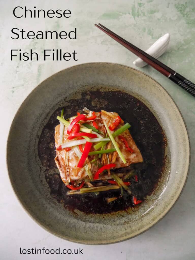 Pinnable image with recipe title and a bowl of Chinese steamed fish fillet with sliced root ginger, spring onion and red chilli, with a hot soy and sesame sauce poured over.