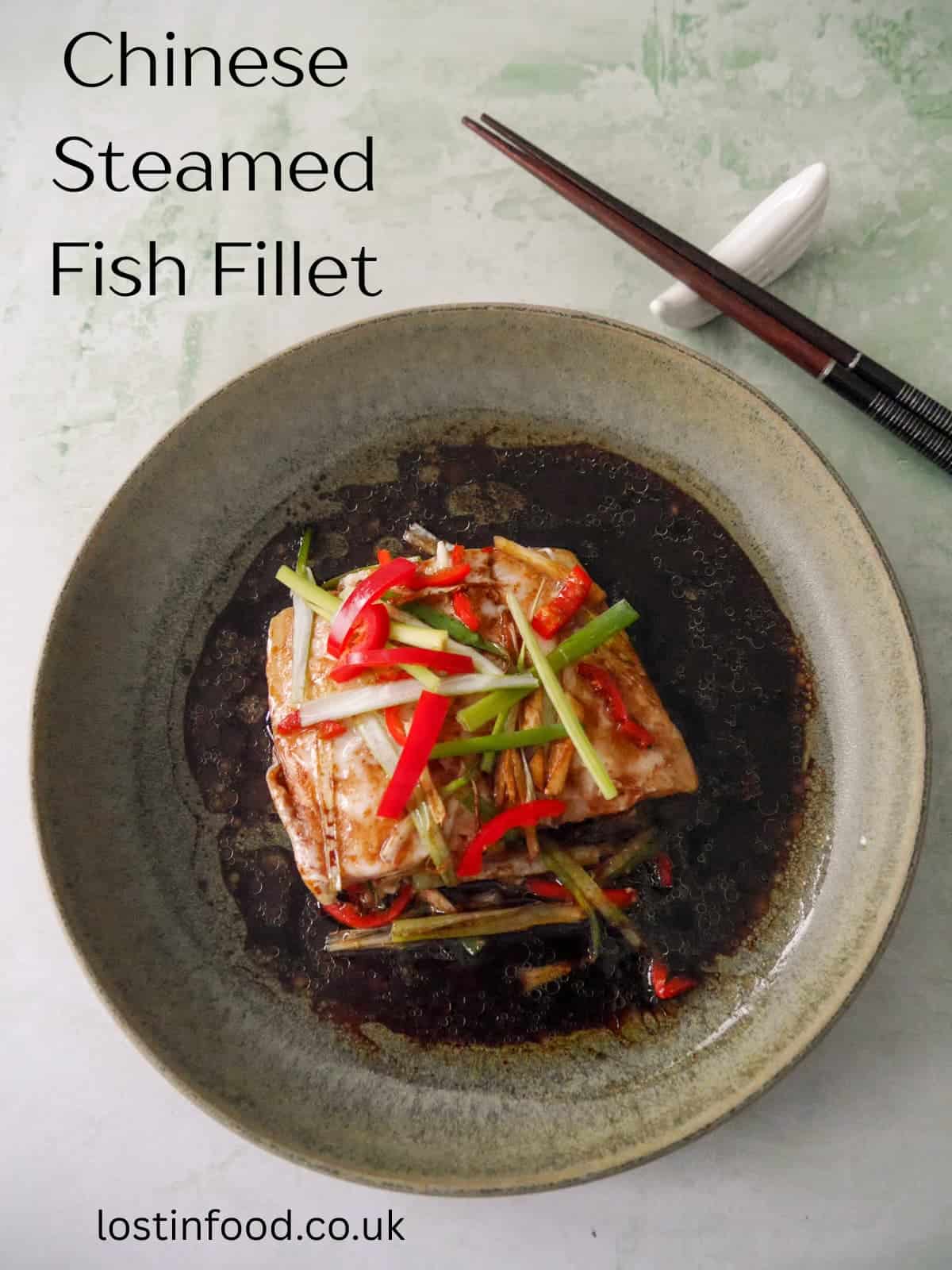 Pinnable image with recipe title and a bowl of Chinese steamed fish fillet with sliced root ginger, spring onion and red chilli, with a hot soy and sesame sauce poured over.