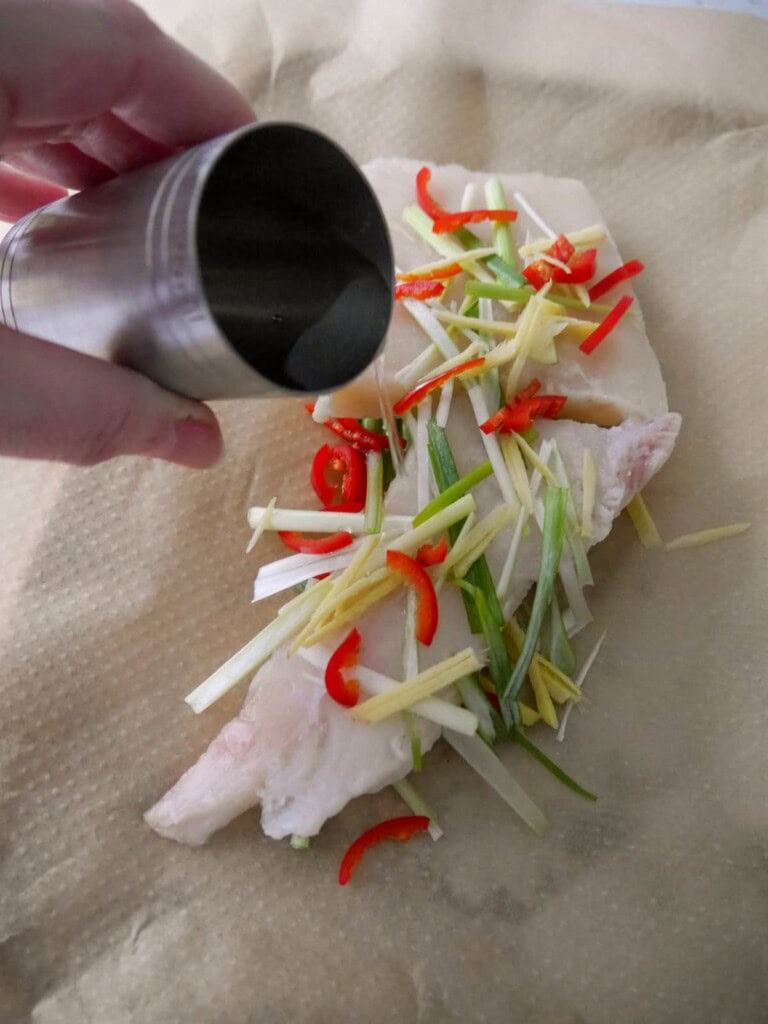 Raw white fish fillets with sliced red chilli, root ginger and spring onions with a splash of water being poured over.