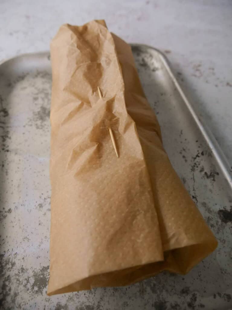 A parchment paper parcel containing fish and aromatics, secured with a cocktail stick and set on a baking tray.