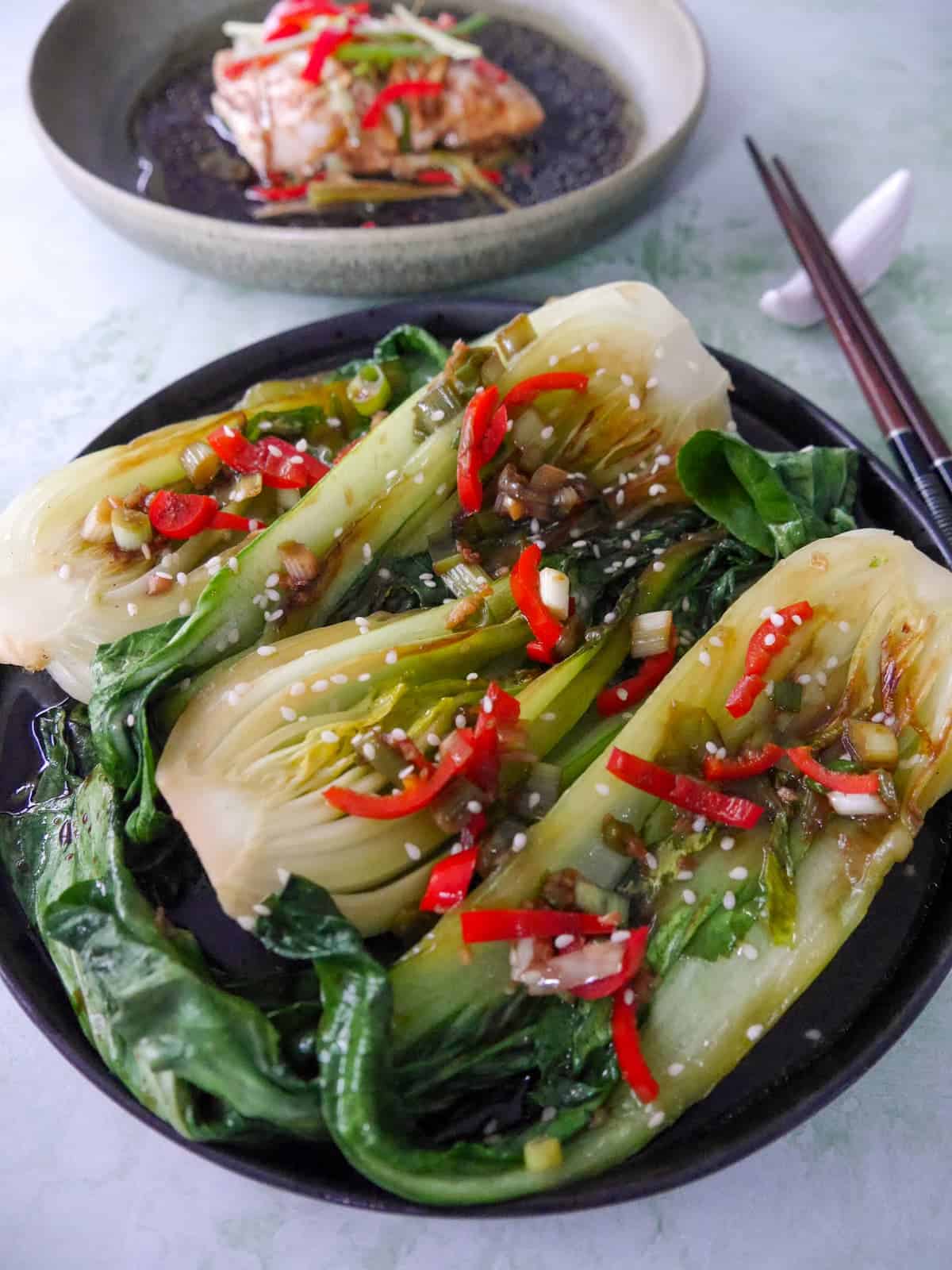 A plate of Chinese stir fry pak choi with garlic, red chillies and spring onion, garnished with sesame seeds, with a bowl of Chinese steamed fish served alongside.