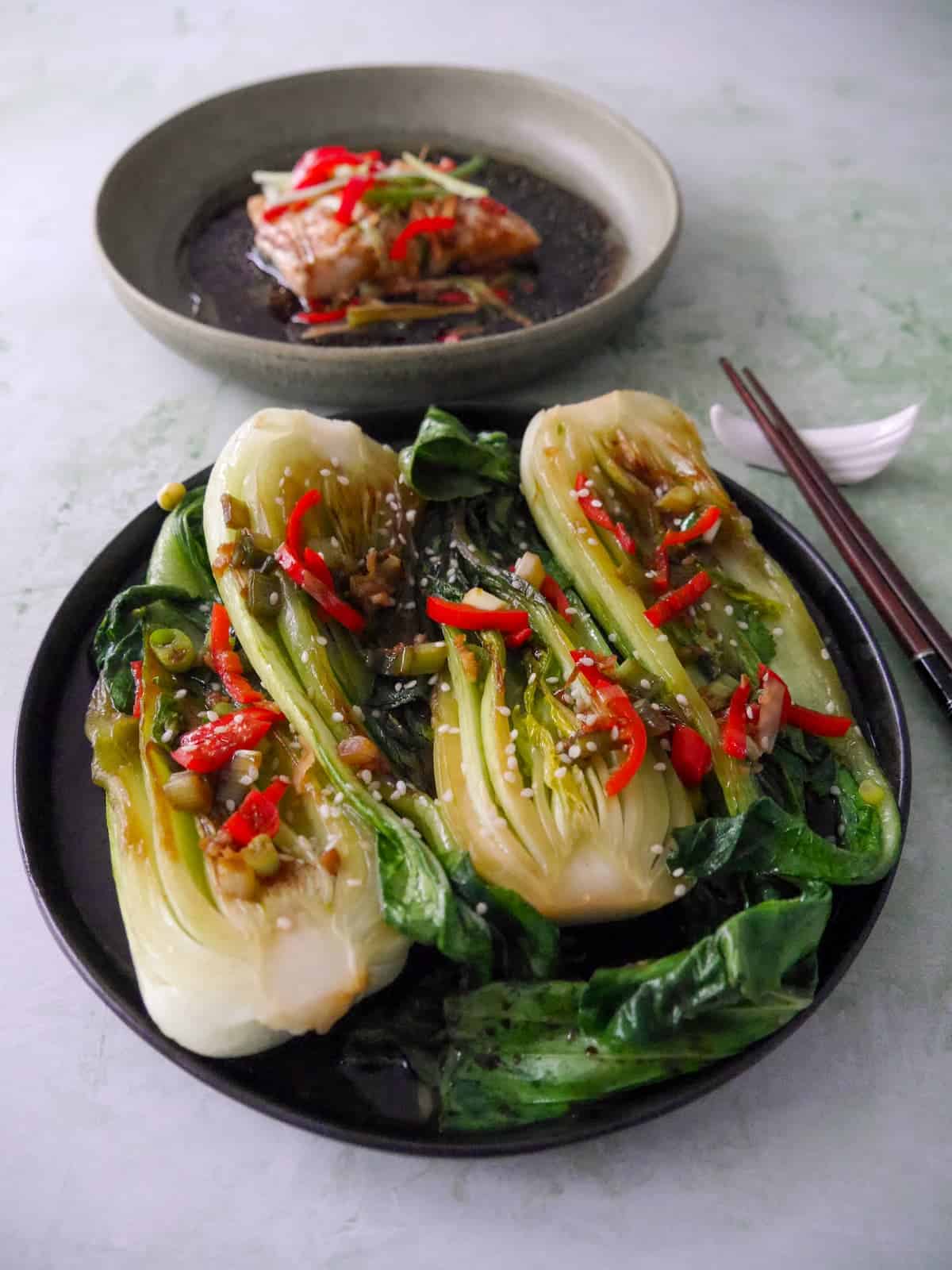 A plate of Chinese stir fry pak choi with garlic, red chillies and spring onion, garnished with sesame seeds, with a bowl of Chinese steamed fish served alongside.