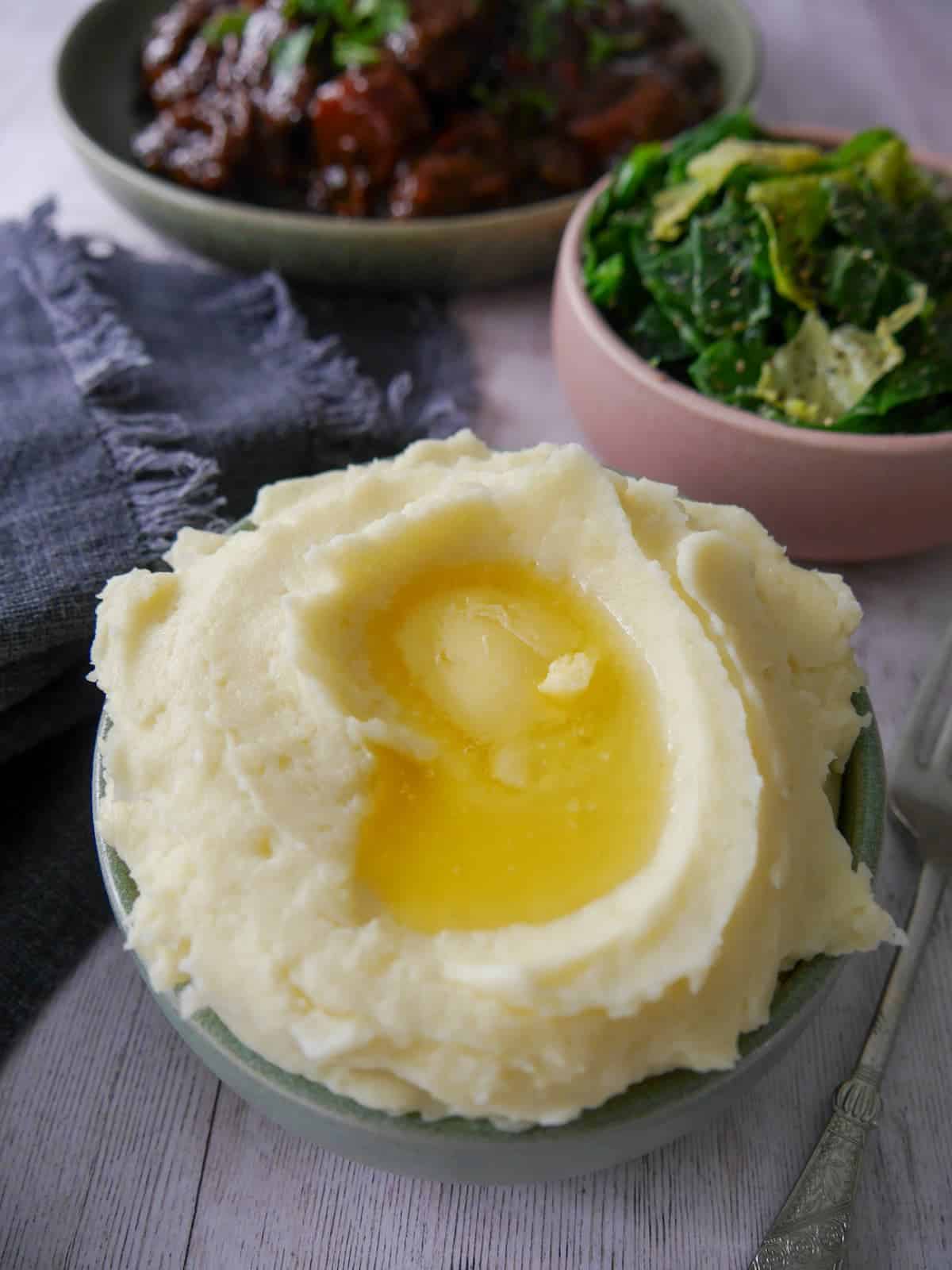 A bowl of creamy mashed potatoes, topped with some butter with a bowl of paprika beef, and a bowl of buttered spring greens set alongside.