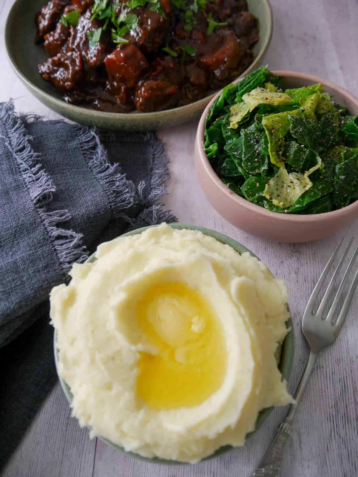 A bowl of creamy mashed potatoes, topped with some butter with a bowl of paprika beef, and a bowl of buttered spring greens set alongside.