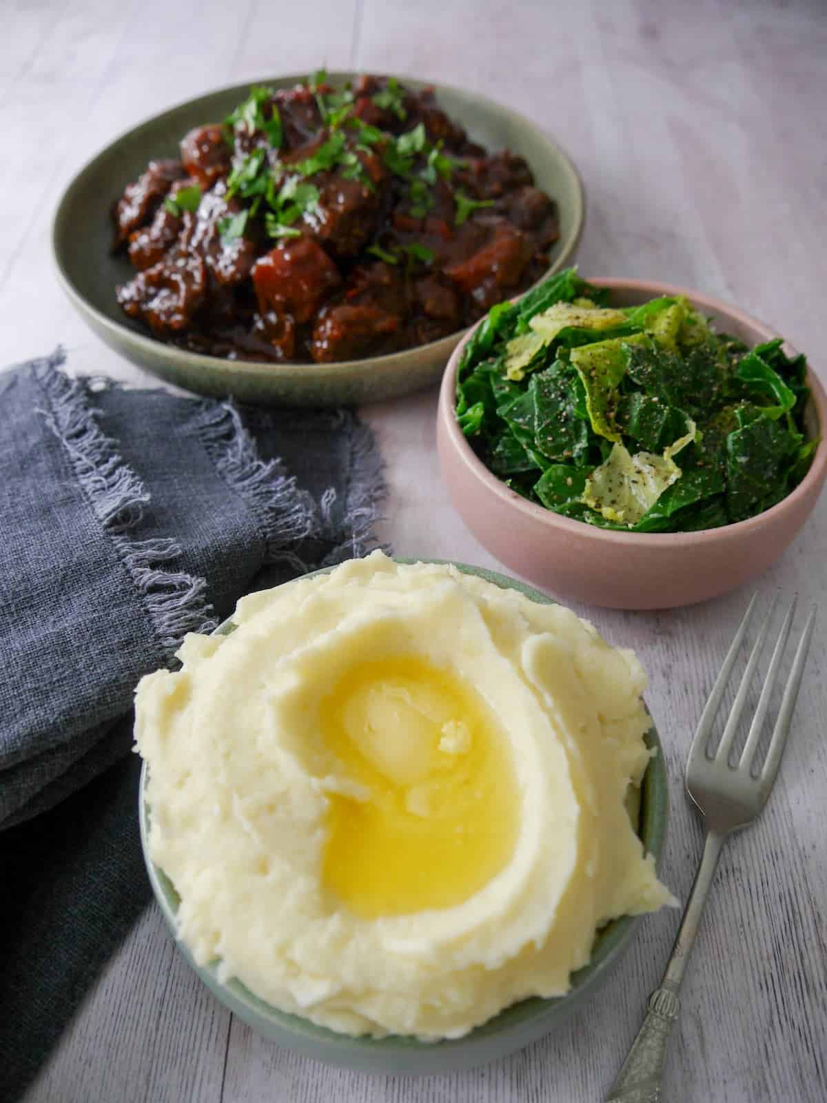 A bowl of creamy mashed potatoes, topped with some butter with a bowl of paprika beef, and a bowl of buttered spring greens set alongside.