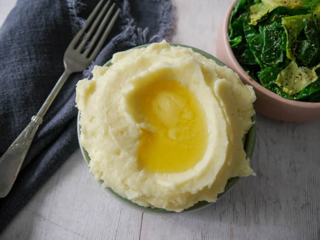 A bowl of creamy mashed potatoes, topped with some butter and a bowl of buttered spring greens set alongside.