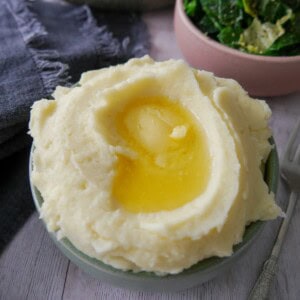 A bowl of creamy mashed potatoes, topped with some butter and a bowl of buttered spring greens set alongside.