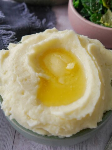 A bowl of creamy mashed potatoes, topped with some butter and a bowl of buttered spring greens set alongside.