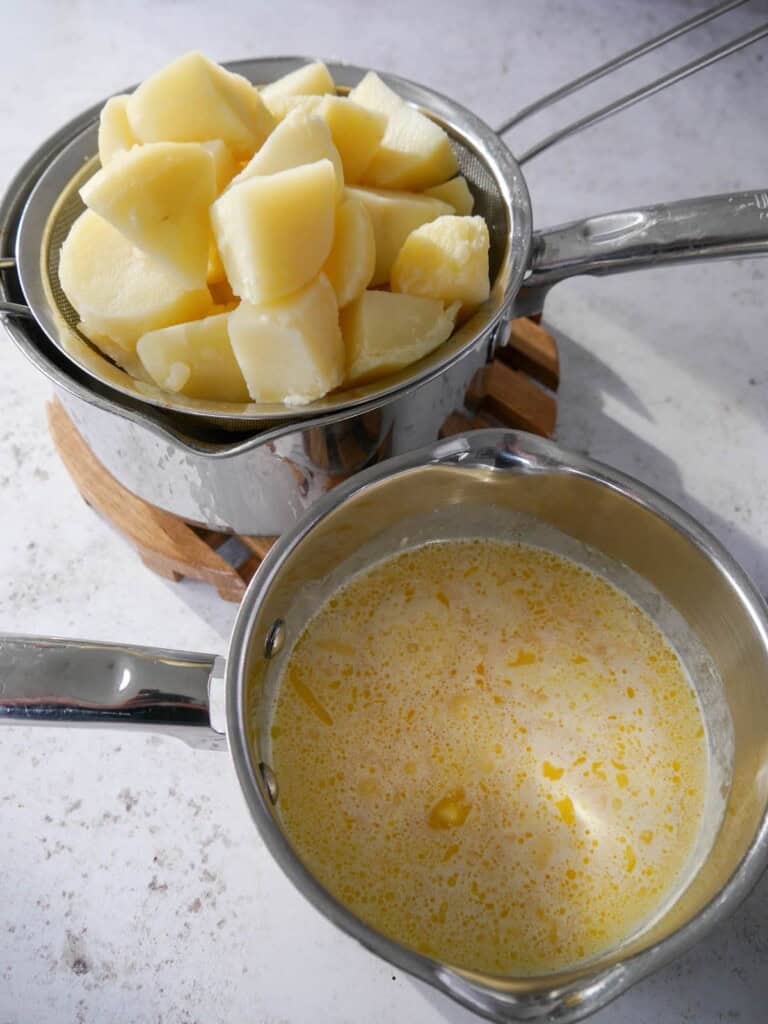 A sieve with cooked boiled potatoes with a small saucepan of warmed butter and cream set alongside.