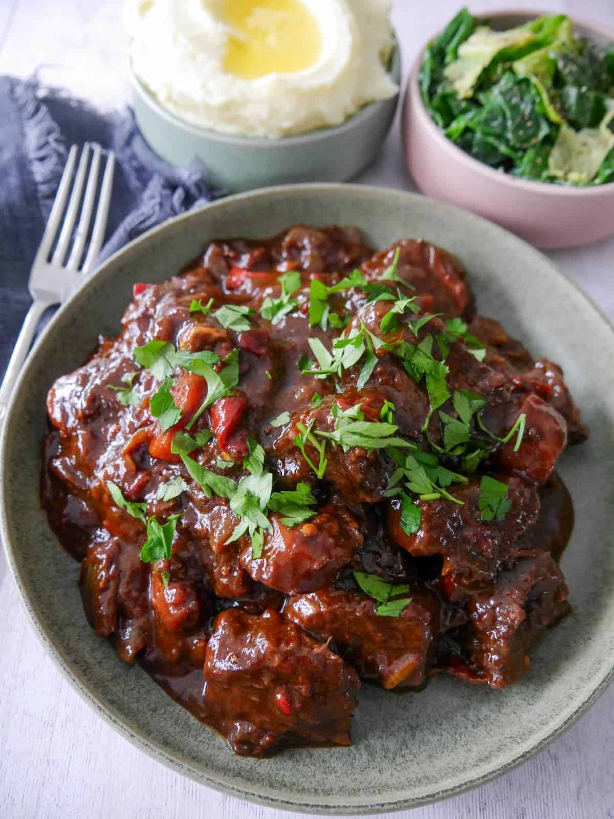 A bowl of paprika beef garnished with chopped parsley, with bowls of creamy mashed potato and buttered spring greens set alongside.