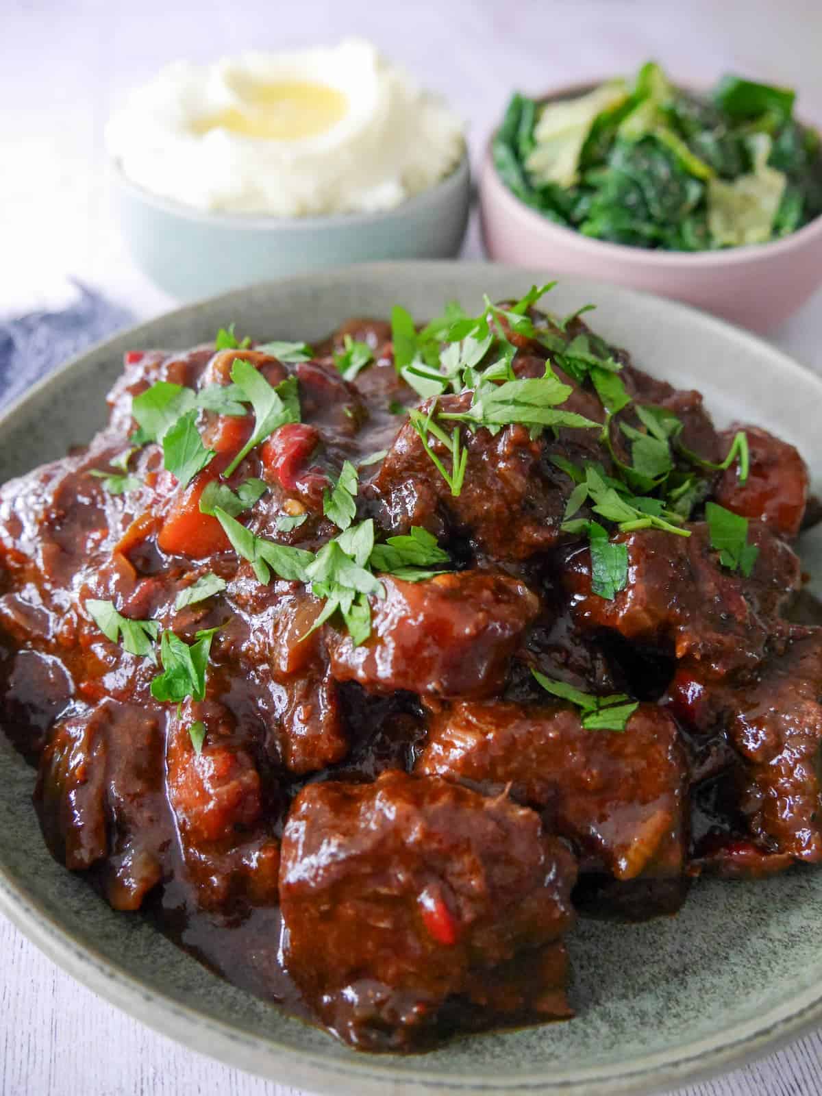 A bowl of paprika beef garnished with chopped parsley, with bowls of creamy mashed potato and buttered spring greens set alongside.