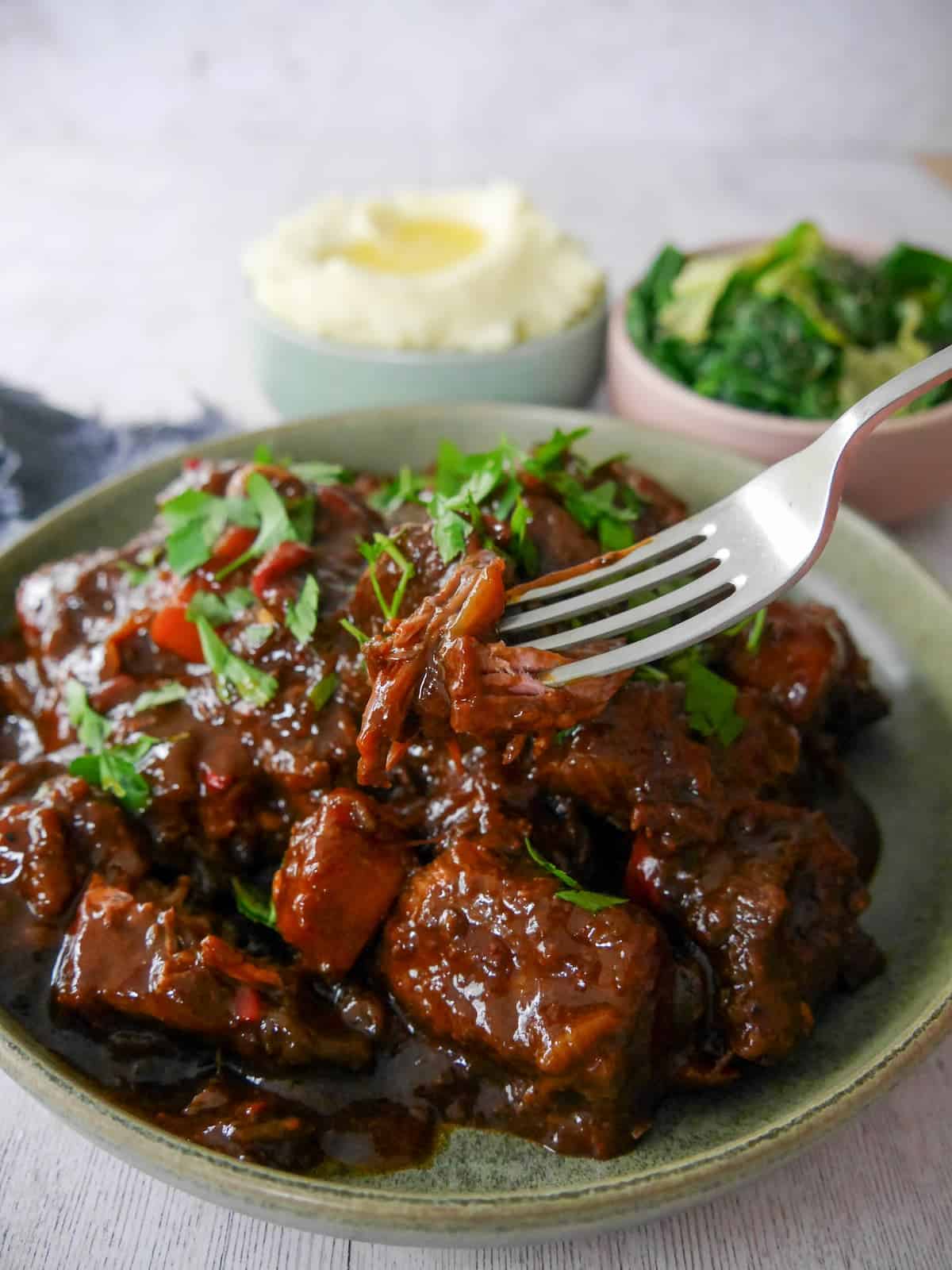 A bowl of paprika beef garnished with chopped parsley, with bowls of creamy mashed potato and buttered spring greens set alongside.