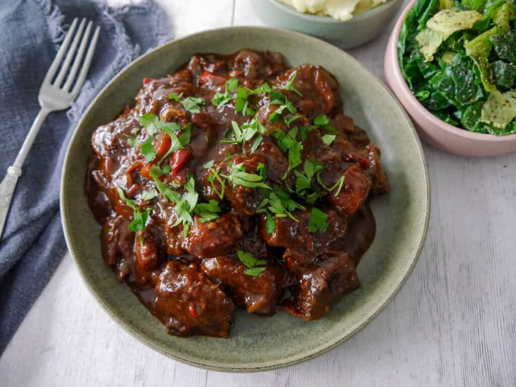 A bowl of paprika beef garnished with chopped parsley, with bowls of creamy mashed potato and buttered spring greens set alongside.