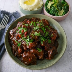 A bowl of paprika beef garnished with chopped parsley, with bowls of creamy mashed potato and buttered spring greens set alongside.