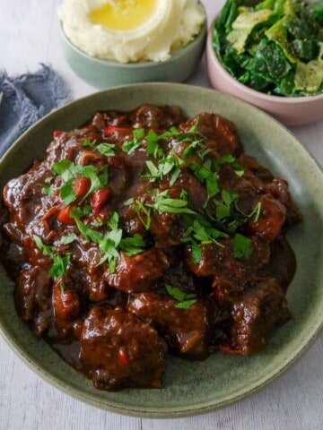 A bowl of paprika beef garnished with chopped parsley, with bowls of creamy mashed potato and buttered spring greens set alongside.