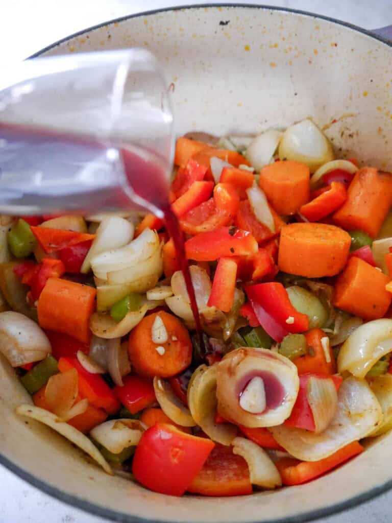 A Dutch oven filled with chunky sauteed vegetables with red wine being poured over.