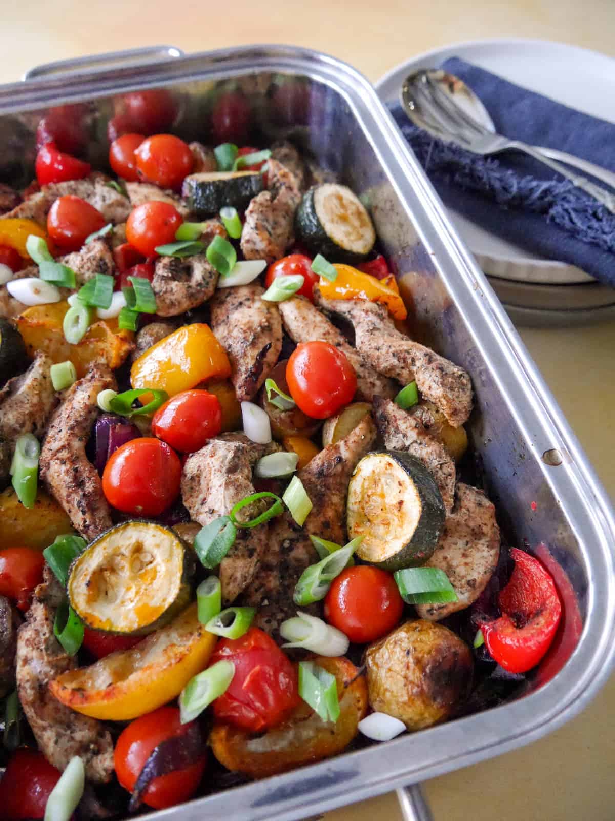 A baking tray filled with roasted vegetables, lemon wedges and topped with strips of cooked Cajun spiced chicken.
