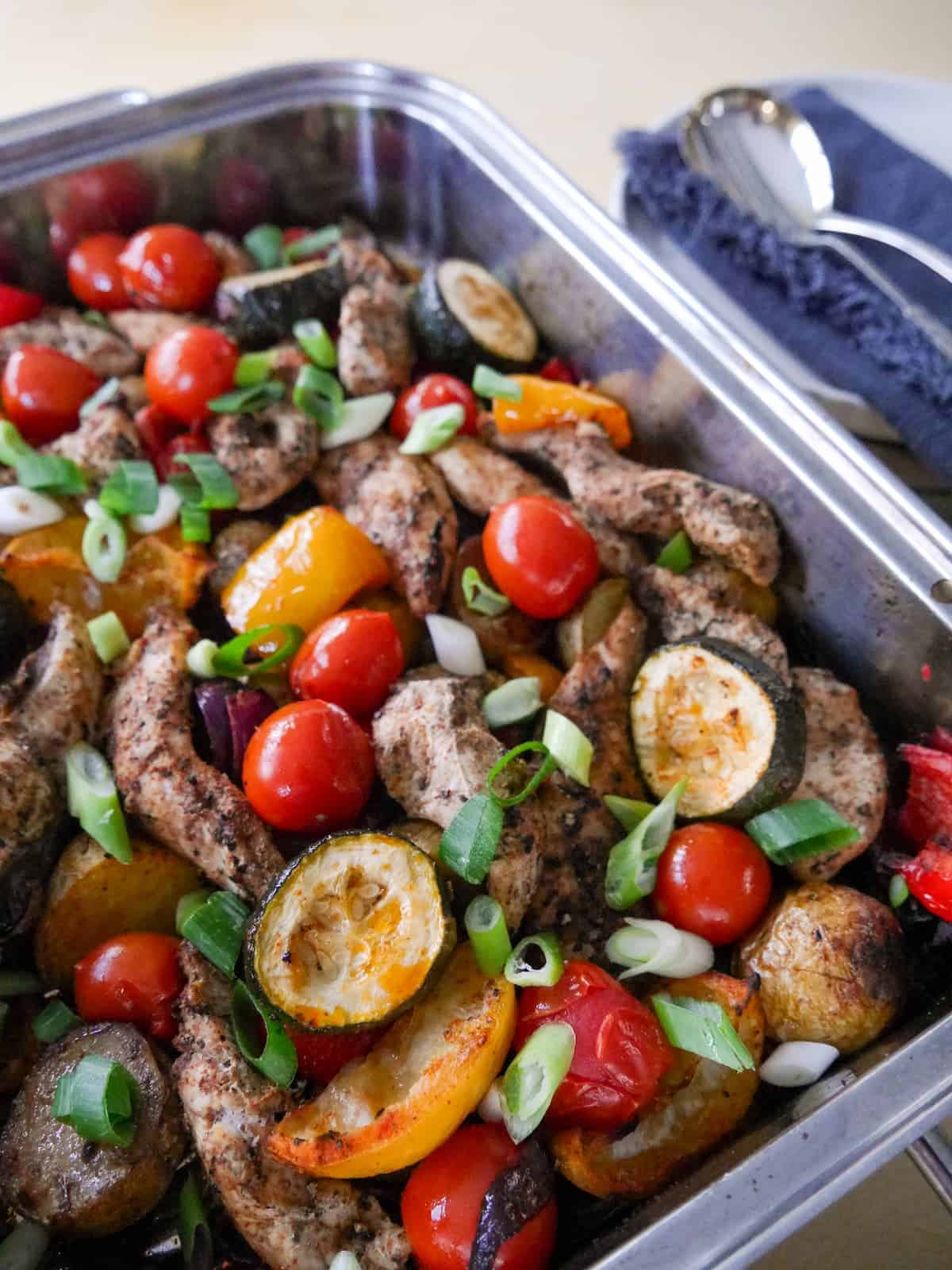 A baking tray filled with roasted vegetables, lemon wedges and topped with strips of cooked Cajun spiced chicken.