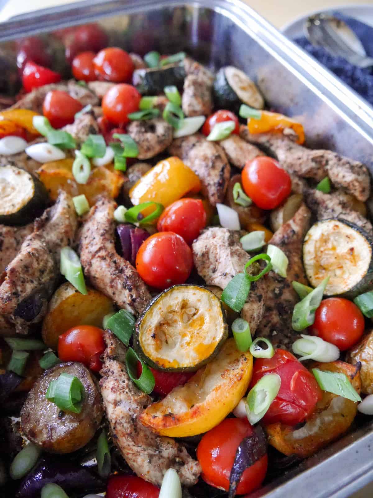 Close up image of a baking tray filled with roasted vegetables, lemon wedges and topped with strips of cooked Cajun spiced chicken.