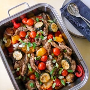 A baking tray filled with roasted vegetables, lemon wedges and topped with strips of cooked Cajun spiced chicken.