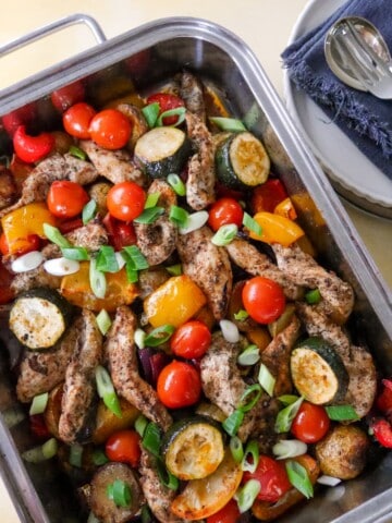 A baking tray filled with roasted vegetables, lemon wedges and topped with strips of cooked Cajun spiced chicken.