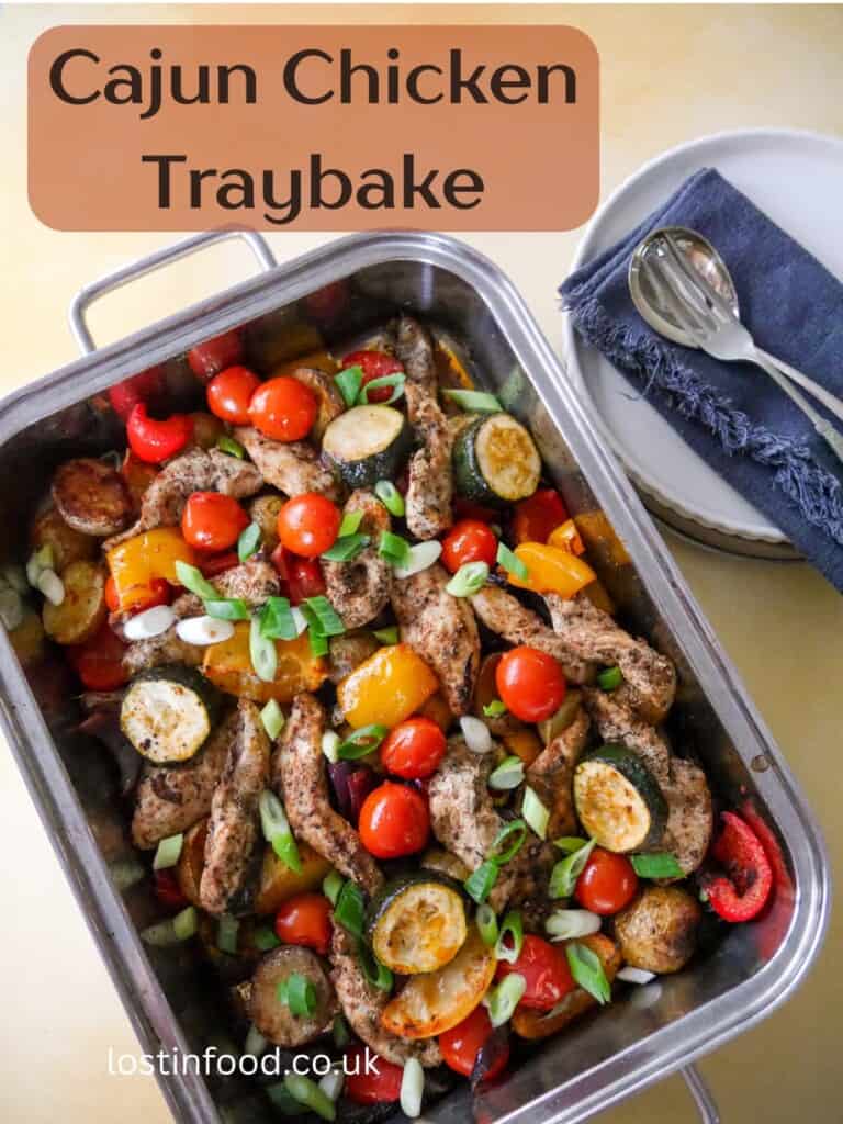 Pinnable image with recipe title and a baking tray filled with roasted vegetables, lemon wedges and topped with strips of cooked Cajun spiced chicken.
