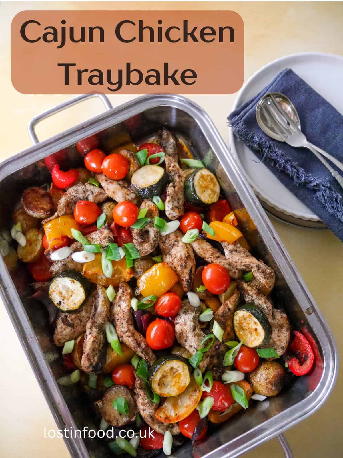 Pinnable image with recipe title and a baking tray filled with roasted vegetables, lemon wedges and topped with strips of cooked Cajun spiced chicken.