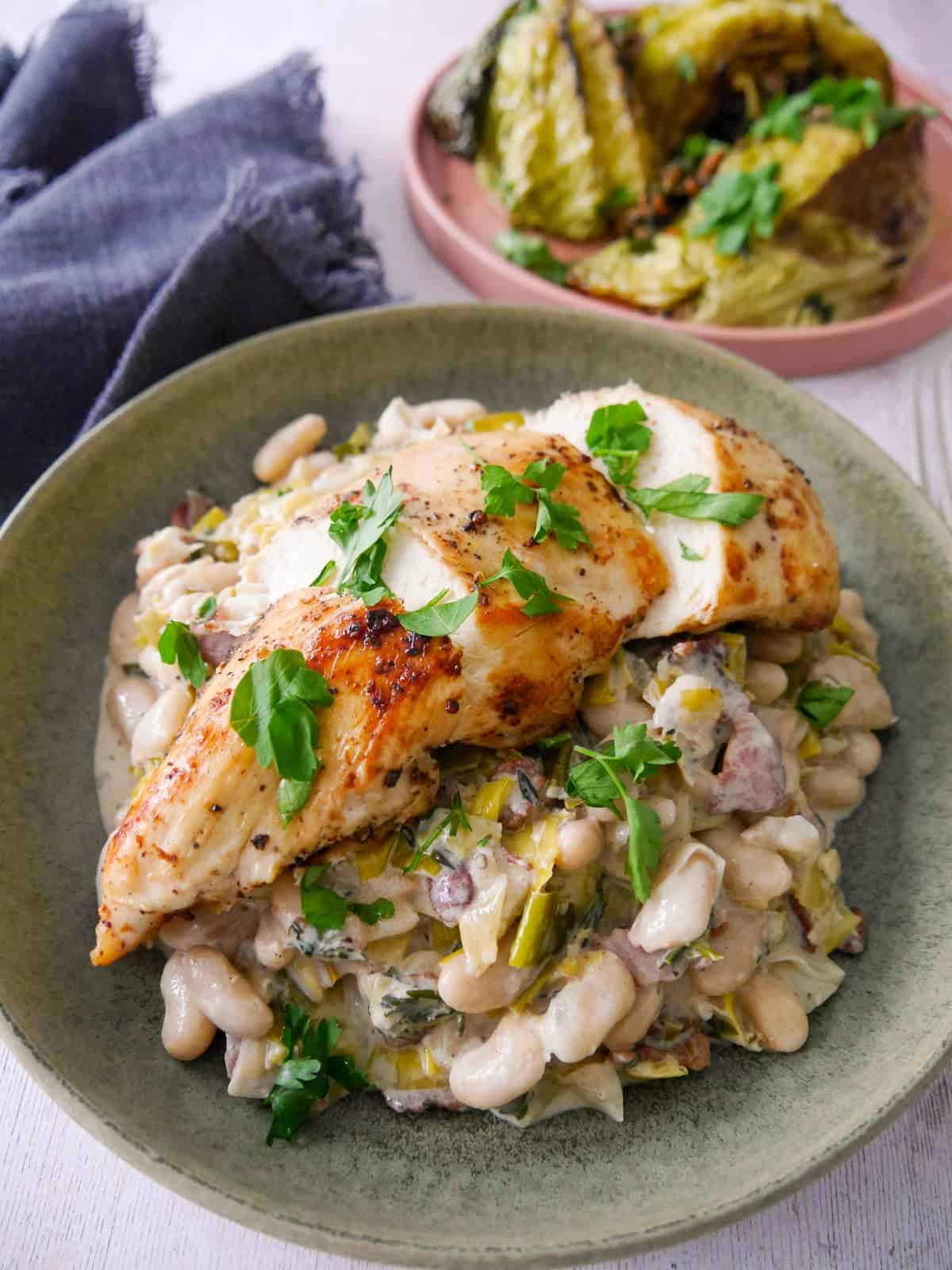 A bowl of cannellini bean stew with bacon and leek topped with sliced cooked chicken breast, with charred cabbage wedges served in a plate alongside.
