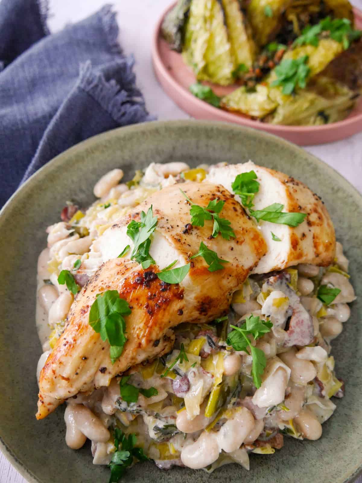 A bowl of cannellini bean stew with bacon and leek topped with sliced cooked chicken breast, with charred cabbage wedges served in a plate alongside.