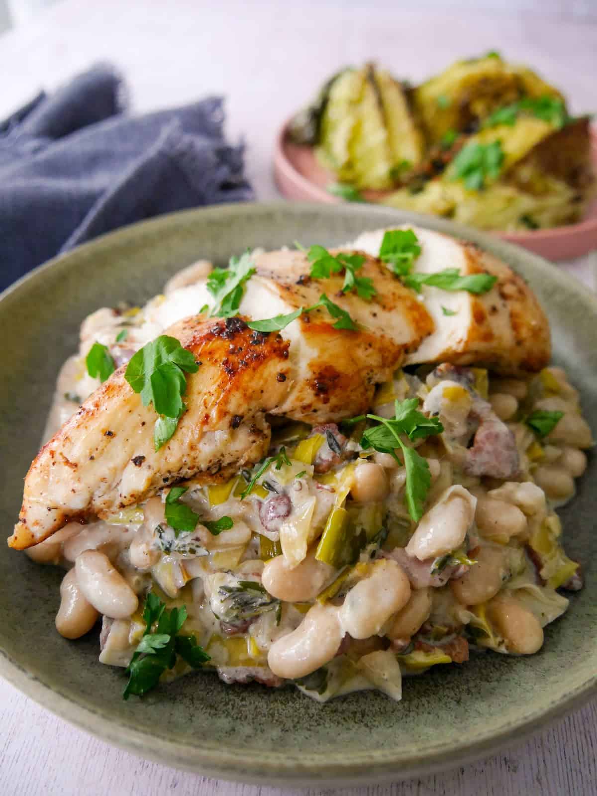 A bowl of cannellini bean stew with bacon and leek topped with sliced cooked chicken breast, with charred cabbage wedges served in a plate alongside.