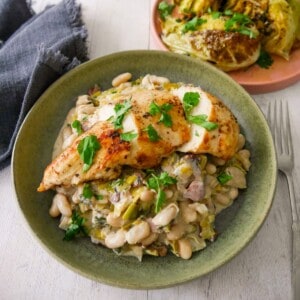 A bowl of cannellini bean stew with bacon and leek topped with sliced cooked chicken breast, with charred cabbage wedges served in a plate alongside.
