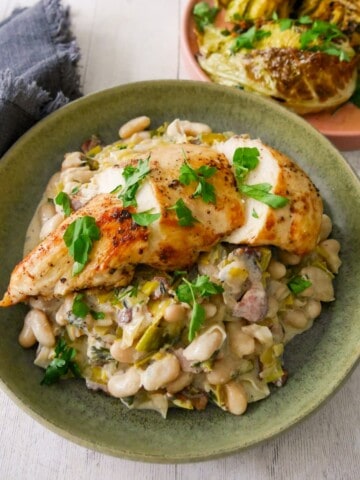A bowl of cannellini bean stew with bacon and leek topped with sliced cooked chicken breast, with charred cabbage wedges served in a plate alongside.