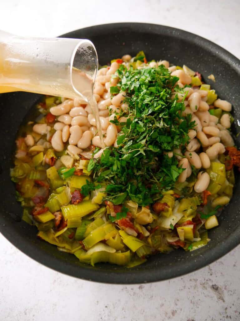 A non-stick frying pan of sauteed bacon and leek with added cannellini beans and fresh herbs with chicken stock being poured over.