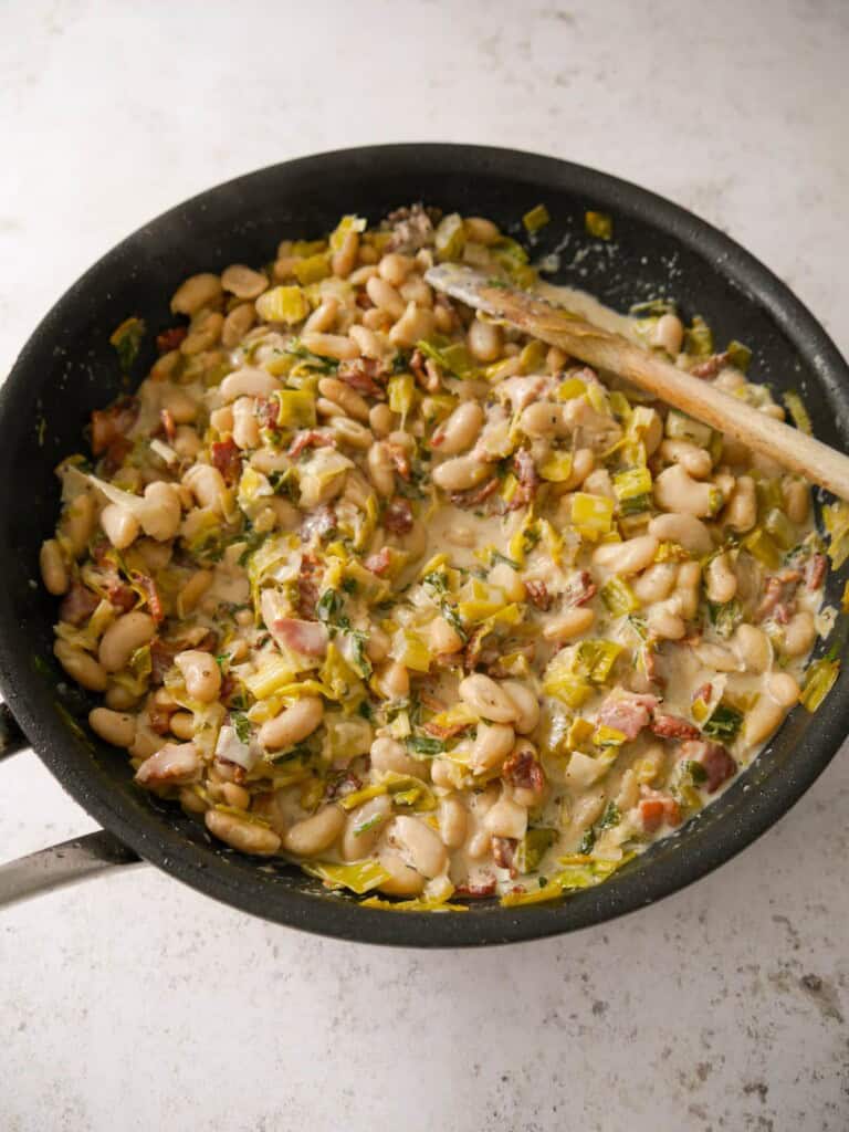 A large non-stick pan of cannellini bean stew with bacon, leeks and fresh herbs.