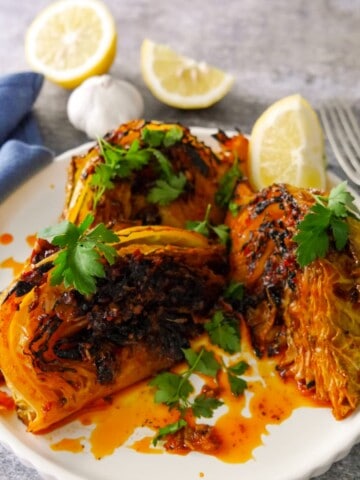 A plate of 3 charred wedges of savoy cabbage cooked in a harissa and lemon butter, garnished with fresh parsley and serve with a wedge of lemon.