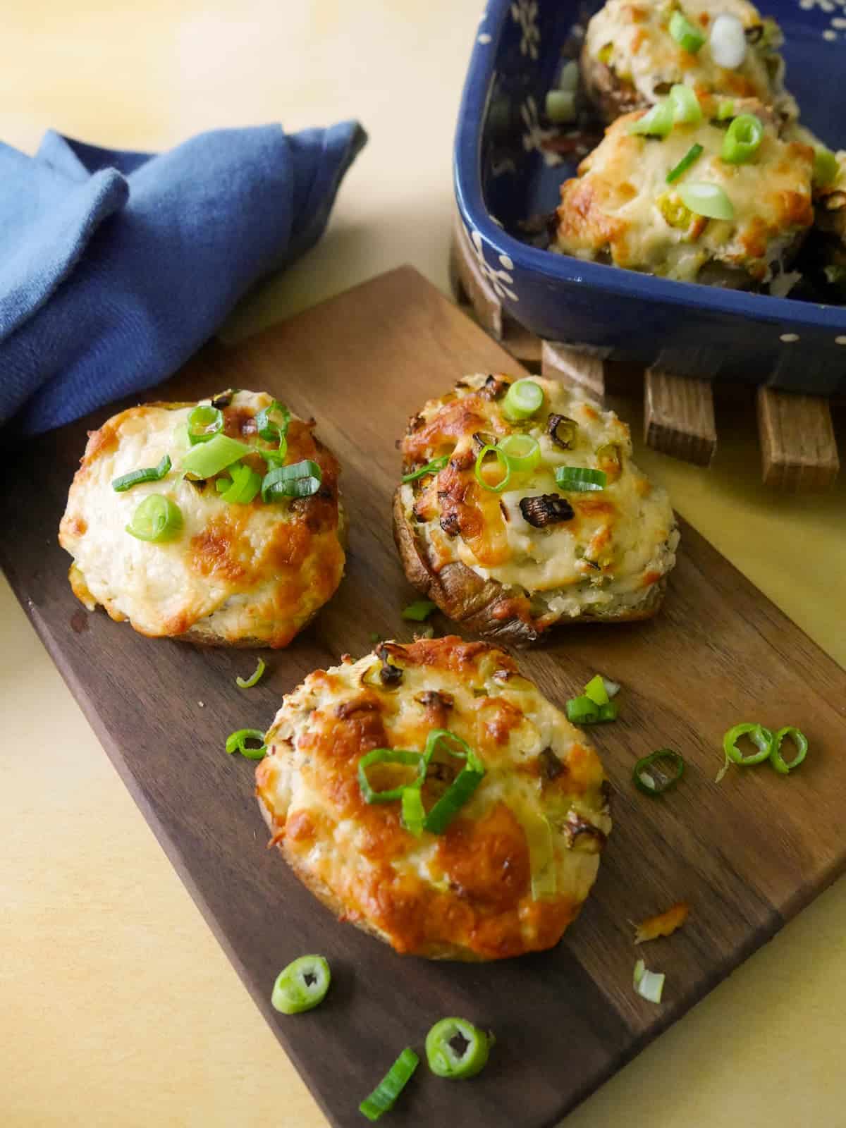 A wooden board topped with cheesy potato skins with a melted cheese and spring onion topping, with an oven proof dish of filled potato skins set alongside.