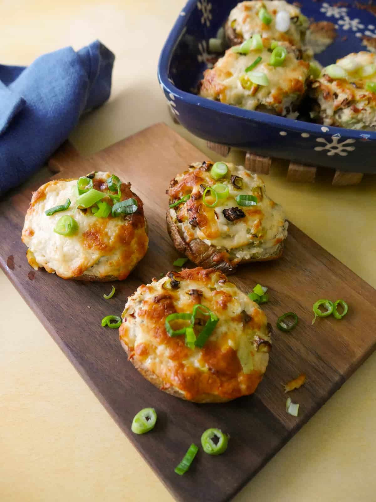 A wooden board topped with cheesy potato skins with a melted cheese and spring onion topping, with an oven proof dish of filled potato skins set alongside.