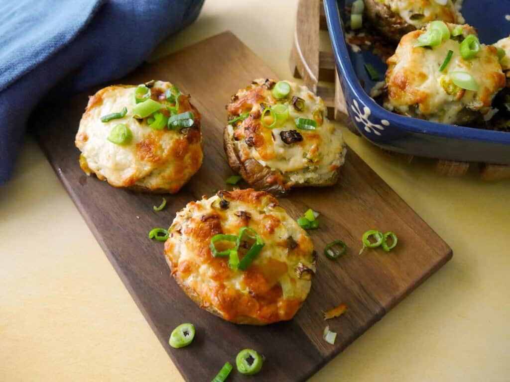 A wooden board topped with cheesy potato skins with a melted cheese and spring onion topping, with an oven proof dish of filled potato skins set alongside.