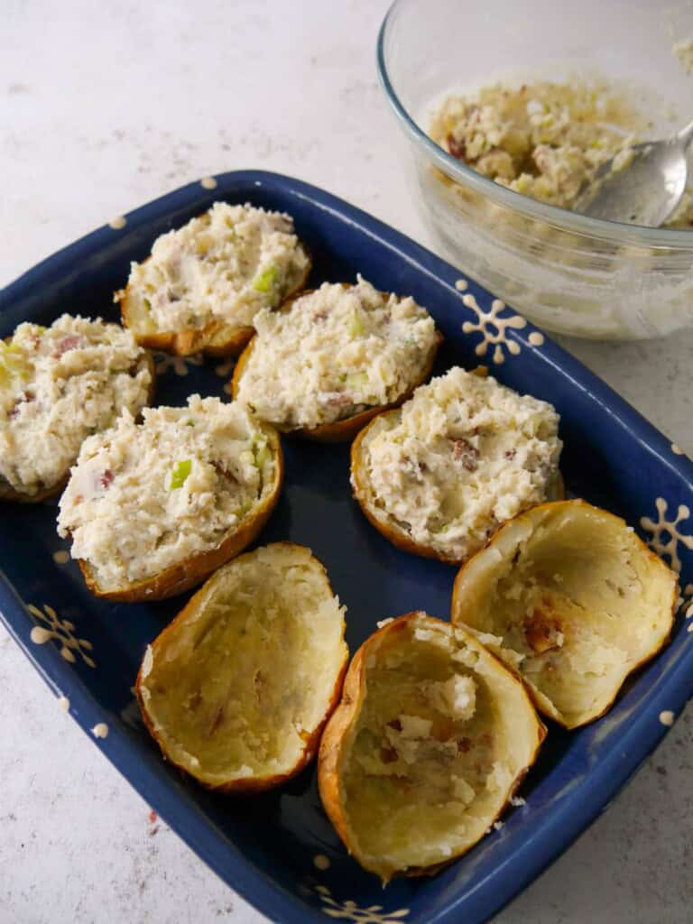 An oven proof dish of halved potatoes, with the flesh scooped out, being filled with a mixture of mashed potato, cream cheese, crispy bacon pieces and spring onions, with a bowl of the cheesy potato mixture set alongside.