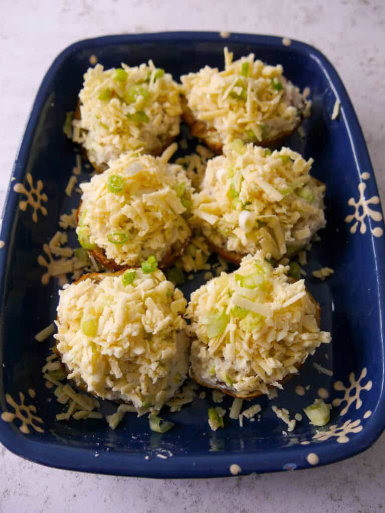 An oven proof dish with cheesy potato skins topped with grated cheddar cheese and sliced spring onions, ready for the oven.