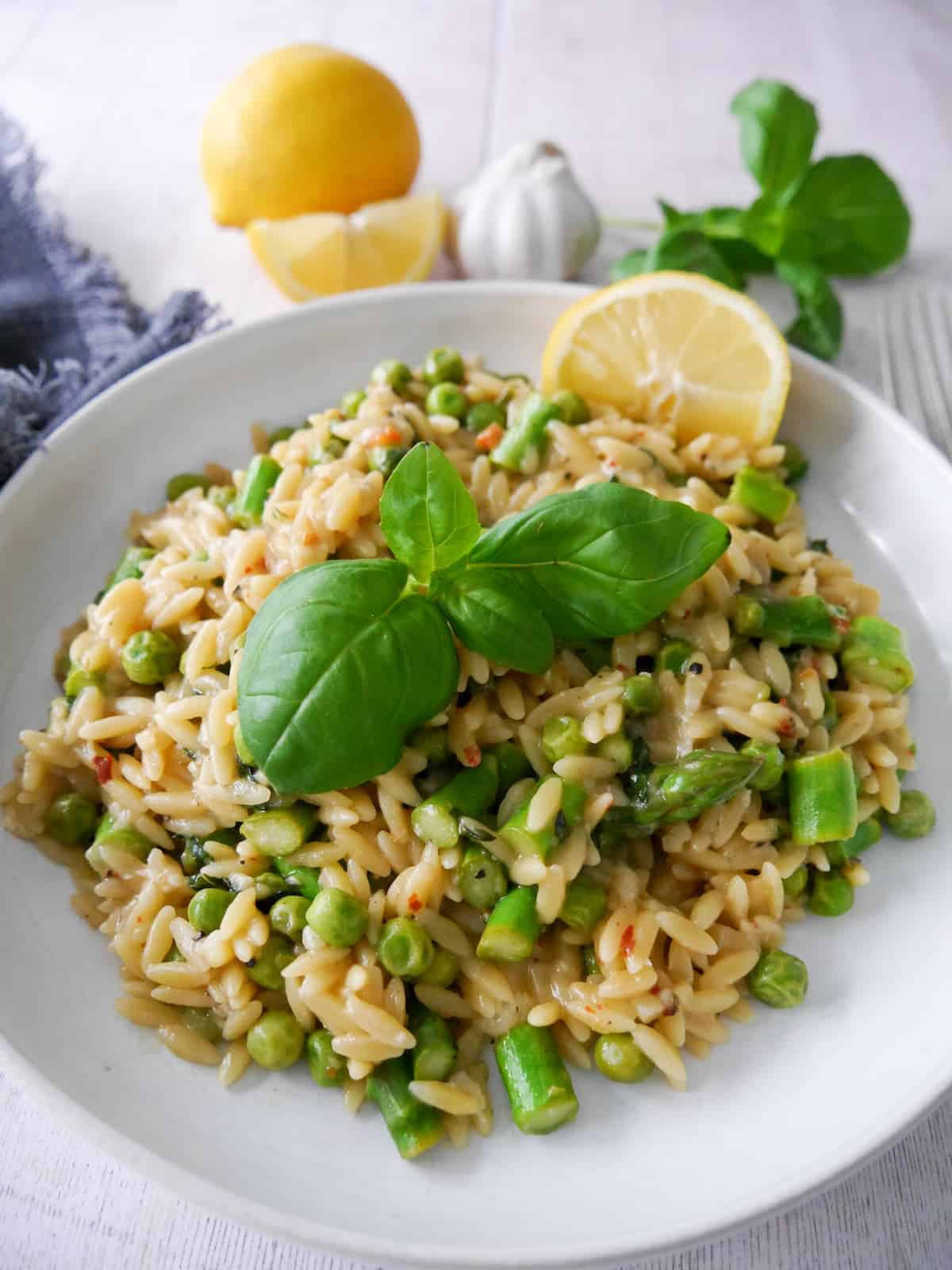 A white bowl of lemon orzo with asparagus and garden peas, garnished with fresh basil and served with a wedge of lemon.