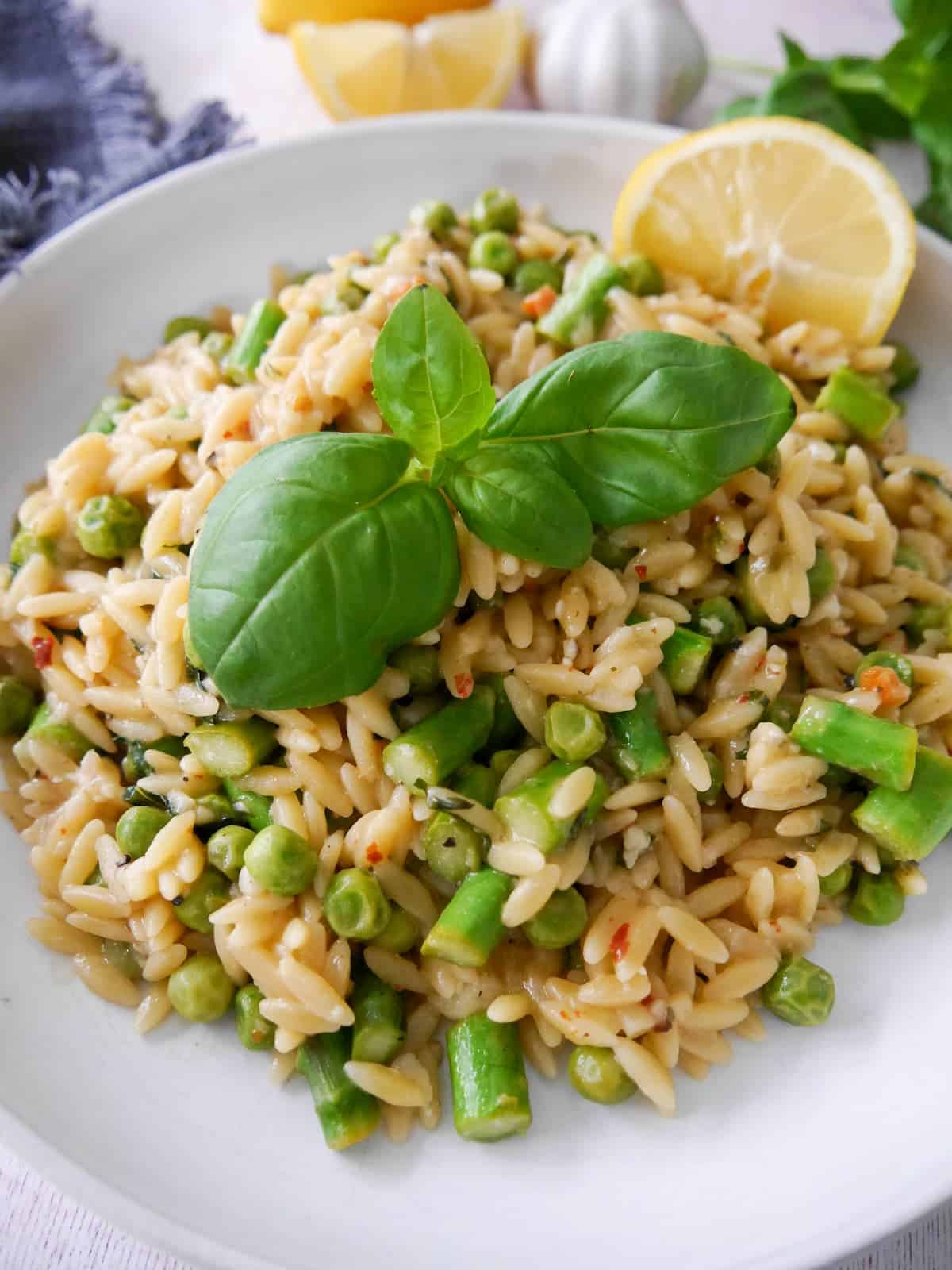 A white bowl of lemon orzo with asparagus and garden peas, garnished with fresh basil and served with a wedge of lemon.