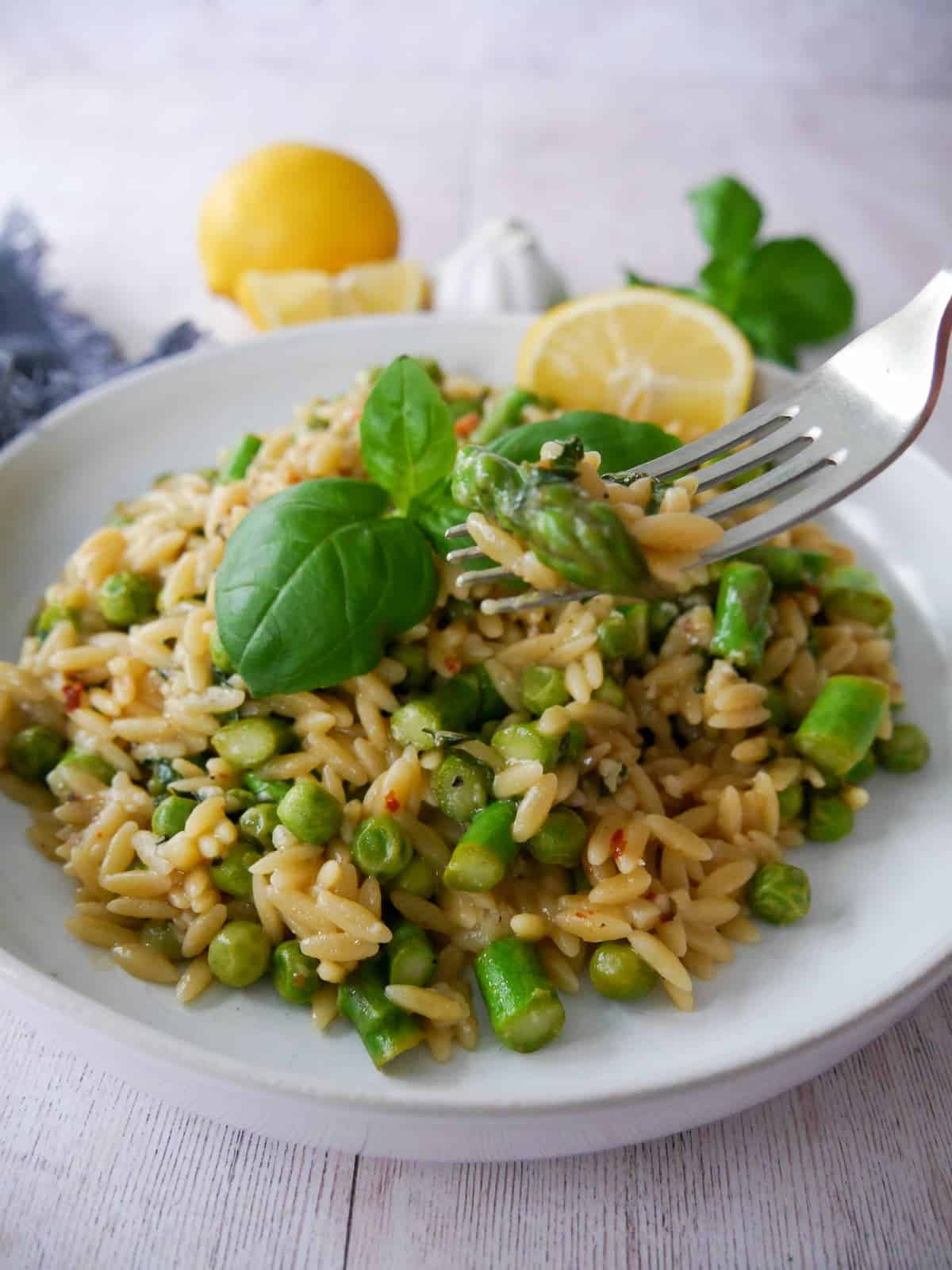 A white bowl of lemon orzo with asparagus and garden peas, garnished with fresh basil and served with a wedge of lemon.